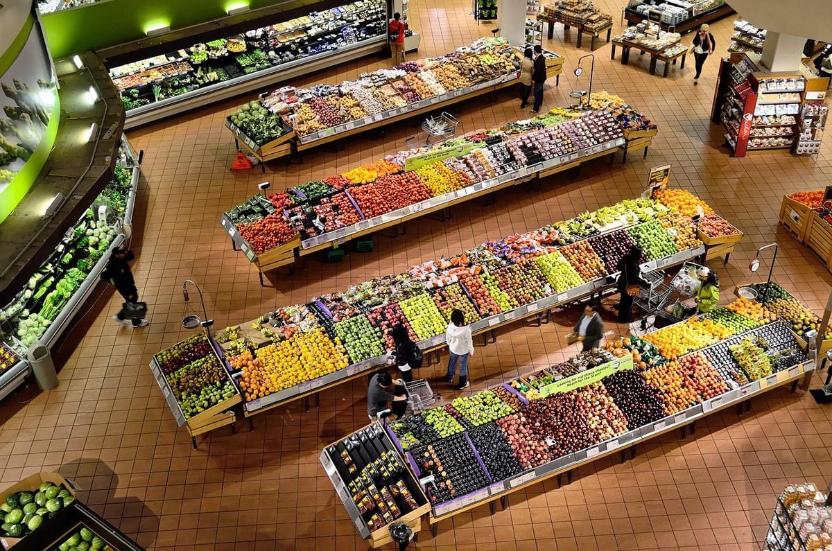 Interior de um supermercado amplo e climatizado, com corredores de frutas e hortifruti